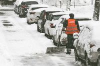 Ein Arbeiter der Firma Garten- und Landschaftsbau in Witten und Umgebung in einer Warnweste räumt Schnee von einer städtischen Straße, während geparkte Autos mit einer dicken Schneeschicht bedeckt sind